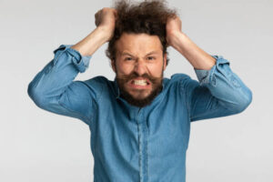 angry bearded man in denim shirt being nervous and pulling hair out, making upset faces and grimacing on grey background in studio