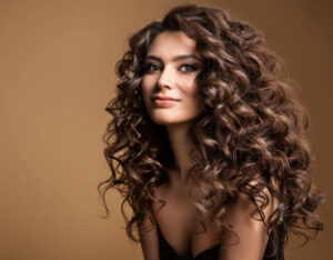 Curly Hair Model. Woman Wavy Long Hairstyle. Brunette Fashion Girl with Volume Hairdo and Natural Make up over Beige Studio Background