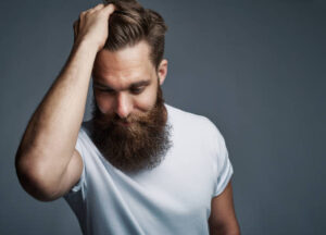 Thinking young man with long beard and white shirt holding hair while facing downward over gray background with copy space